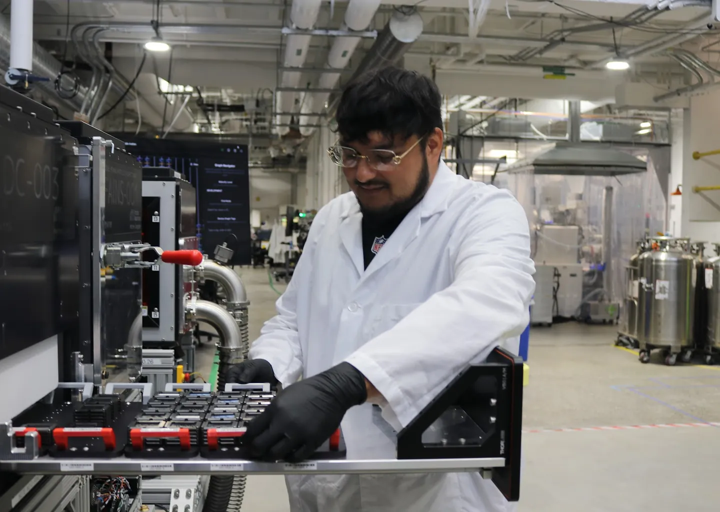 A person in a white lab coat and safety glasses sliding a tray of parts out of a machine on the production floor