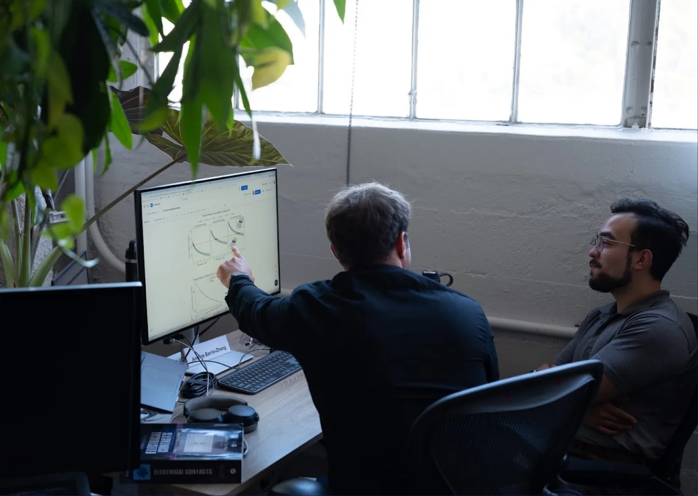 Two people at a desk under a window, one pointing at a graph on a monitor