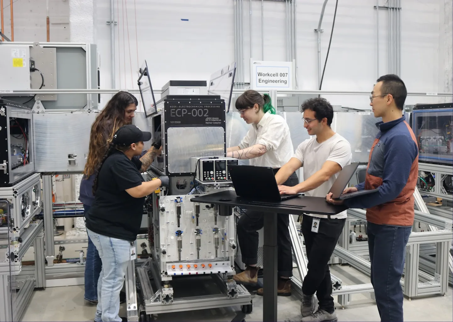 Five people in an engineering workcell, two at a machine and three around a standing desk with laptops